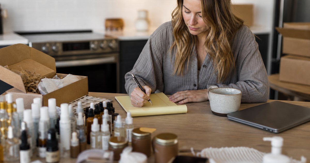 A young entrepreneur at a kitchen table surrounded by sample products and shipping supplies, sorting the samples into two small groups on the table, closed laptop beside a mug