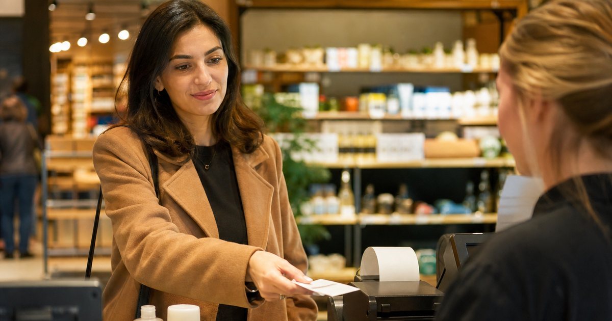 A shopper at a retail checkout counter placing a few small items on the counter while the cashier reaches for them, warm store lighting