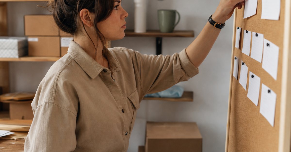 A small business owner standing at a corkboard in a back office, pulling one pushpin from a row of pins holding small fabric swatches, coffee mug and boxes of inventory on a shelf behind